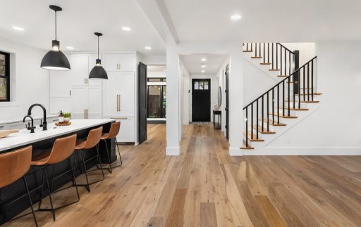 A modern open concept interior features light hardwood flooring, a bright kitchen with an island, and a staircase with black railings.